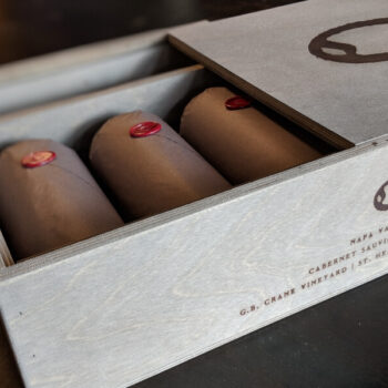 G.B. Crane Vineyard Cabernet Sauvignon 2017 OWC 3 Pack; Rectangular label , dark brown merging with black. Features a grey box, with the lid pulled back revealing three bottles wrapped in brown paper, with round, red wax seals