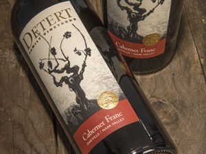 Detert Cabernet Franc 2014 3 Liter; A rectangular label showing two bottles of wine, one standing, one lying down, on a dark wood surface. The bottles have a label that is beige featuring a dark brown grape vine. Text is gold, and gold on deep red.